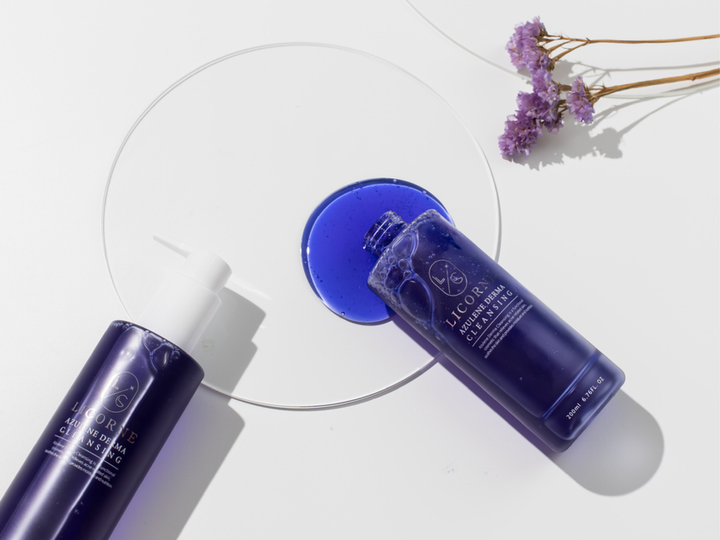 Two blue skincare bottles with a circular mirror and purple flowers on a white background